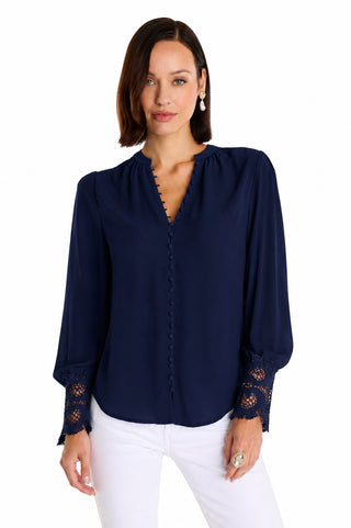 A woman with shoulder-length brown hair models the Blake Lace Sleeve Blouse in navy, featuring long sleeves with lace-detailed cuffs and a high-low hem, paired with white pants against a plain white background.