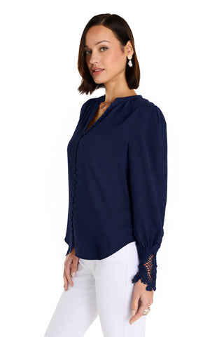 A woman stands sideways and looks at the camera, modeling the Blake Lace Sleeve Blouse in navy with lace-detailed cuffs and a high-low hem, paired with white pants against a white background.