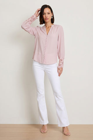 A woman stands against a plain background in the Blake Lace Sleeve Blouse - Blush, white flare pants, nude heeled sandals, and pearl earrings. One hand touches her hair as the other rests at her side.