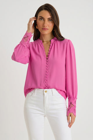 A woman in the Blake Lace Sleeve Blouse in petal pink, featuring decorative buttons and lace cuffs, paired with high-waisted white pants, stands against a plain light background and touches her hair.