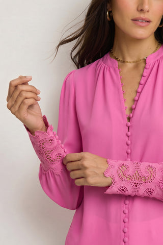 A woman with long brown hair, gold hoop earrings, and a gold necklace gently touches the sleeve of her Blake Lace Sleeve Blouse in Petal Pink, featuring decorative buttons and intricate lace cuffs.