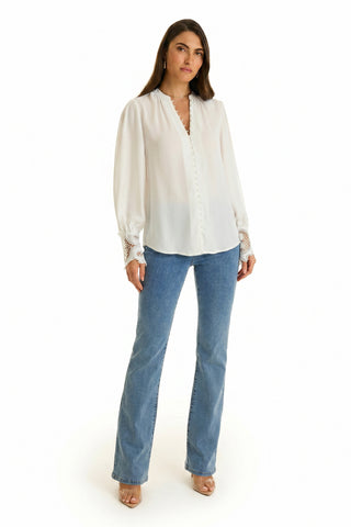 A woman with long brown hair and a neutral expression stands against a white background wearing the Blake Lace Sleeve Blouse in white, light blue bootcut jeans, and nude heels.