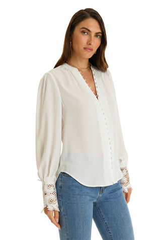 A woman wearing the Blake Lace Sleeve Blouse in white with intricate cuffs and blue jeans stands against a plain white background, looking slightly to the side.