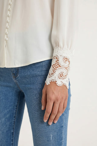 A person wearing light blue jeans and the Blake Lace Sleeve Blouse in white—a feminine long-sleeve top with lace and embroidered cuffs—stands with their arm relaxed by their side.