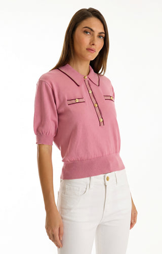 A woman with long brown hair wears the Bobbi Polo Shirt - Dusty Rose, featuring gold buttons, paired with high-waisted white pants for a chic casual look, standing against a plain white background.