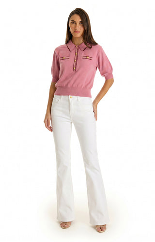 A woman with straight brown hair faces forward, wearing the Bobbi Polo Shirt - Dusty Rose, an ultra-soft knit pink short-sleeve sweater with front pockets, paired with white high-waisted flared pants and open-toed heels.