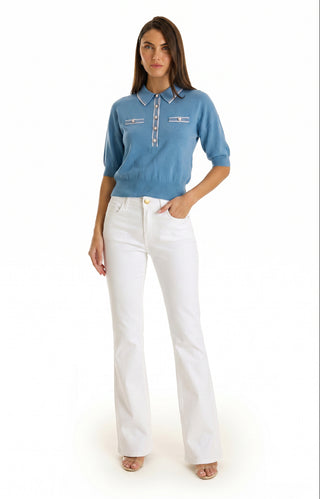 A woman stands facing forward wearing the Bobbi Polo Shirt in Serene Blue with embossed dome buttons and white high-waisted, flared jeans, one hand in her pocket, open-toed shoes, against a plain white background.