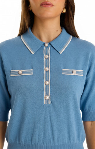 A woman in the Bobbi Polo Shirt - Serene Blue, an ultra-soft knit polo with a collar and dome buttons on the front and two decorative chest pockets. She accessorizes with gold earrings and has straight brown hair.
