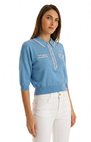 A woman in a Serene Blue Bobbi Polo Shirt with ultra-soft knit, white trim, and embossed dome buttons, paired with high-waisted white pants, stands against a plain white background.