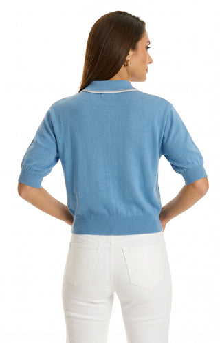 A woman with long brown hair is seen from the back, wearing the Bobbi Polo Shirt - Serene Blue paired with white pants. The ultra-soft knit shirt features a collar and is set against a plain white background.