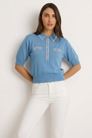 A woman with straight brown hair and a relaxed expression stands against a light background, wearing the Bobbi Polo Shirt in Serene Blue with white trim and buttons, paired with high-waisted white pants.