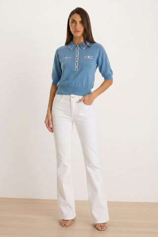 A woman stands indoors wearing the Bobbi Polo Shirt in Serene Blue and white high-waisted flare pants. With straight brown hair, she poses confidently with one hand in her pocket, looking at the camera.