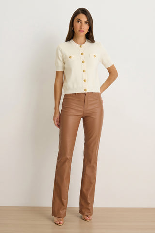 A woman with straight brown hair and a neutral expression stands against a white wall wearing the Bonnie Knit Cardigan Top in Pearl, featuring short sleeves and gold dome buttons. She pairs it with high-waisted brown leather pants and sandals.