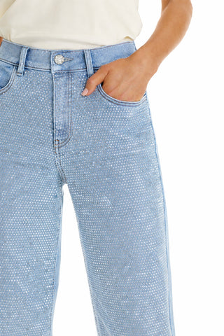 A person models the Brynn Crystal Jean - Medium Blue, featuring a high-rise wide-leg fit with small white polka dots, paired with a white top and one hand in their pocket against a white background.