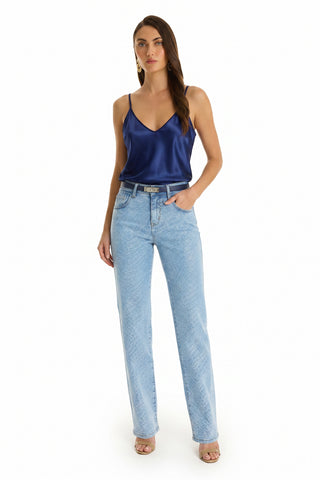 A woman with long brown hair stands against a white background wearing a blue satin camisole, Brynn Crystal Jean - Medium Blue (high-rise, wide-leg with subtle crystal details), beige heeled sandals, and gold hoop earrings. One hand is in her pocket.