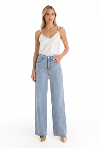 A woman with light hair poses confidently against a white background in a white sleeveless camisole, beige pointed-toe heels, and the Brynn Crystal Jean - Medium Blue, effortlessly highlighting its high-rise wide-leg style.