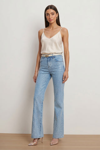 A woman with long brown hair and a neutral expression stands against a plain background wearing a cream camisole, beige belt, clear pointed-toe heels, and the Brynn Crystal Jean in Medium Blue with a subtle print.