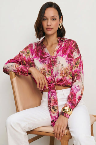 A woman with short brown hair sits on a wooden chair, wearing the Cecilia Silk Tie-Front Blouse in Bright Orchid, white pants, gold earrings, and a chunky gold bracelet, looking confidently at the camera.