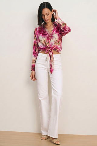 A woman with shoulder-length dark hair stands against a plain background wearing the Cecilia Silk Tie-Front Blouse in Bright Orchid, white high-waisted wide-leg pants, and beige heeled sandals. She looks down, gently touching her hair.