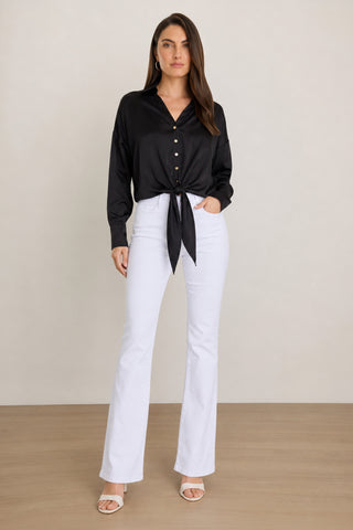 A woman stands indoors against a plain light-colored background, wearing the versatile Cecilia Silk Tie-Front Blouse in black, styled at the waist, paired with white high-waisted flared pants and white sandals.