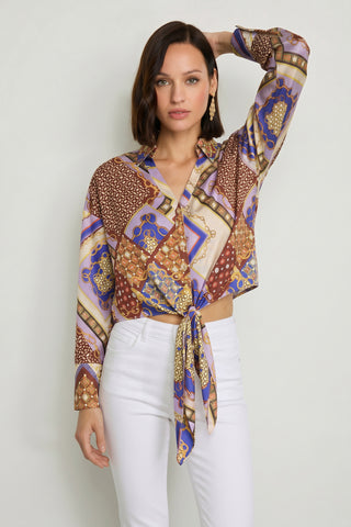 A woman with shoulder-length dark hair wears the Cecilia Silk Tie-Front Blouse in Riviera Chain, styled with white high-waisted pants. She stands against a light background and accessorizes with gold earrings.