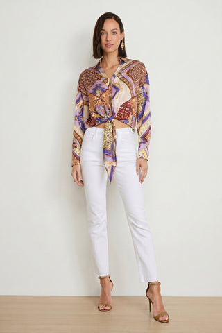 A woman with shoulder-length brown hair and minimal accessories stands against a plain wall wearing the Cecilia Silk Tie-Front Blouse – Riviera Chain, white high-waisted pants, and tan high-heeled sandals.