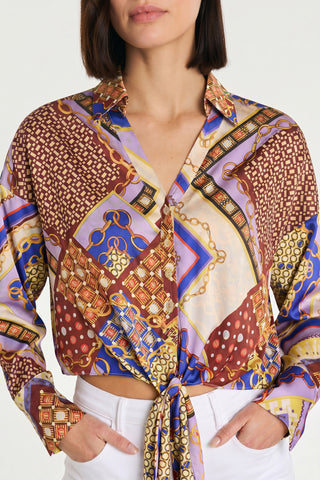 A woman wears the Cecilia Silk Tie-Front Blouse – Riviera Chain, featuring red, blue, gold, and purple geometric and chain prints. She has one hand in her white pants pocket with her face partially visible.