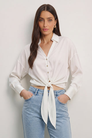 A woman with long brown hair wears the Cecilia Silk Tie-Front Blouse in white, paired with light blue jeans and standing hands-in-pockets against a plain white background—a perfect look for effortless resort style.