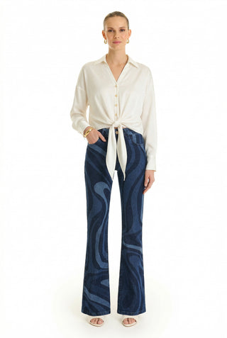 A woman stands facing forward, wearing the Cecilia Silk Tie-Front Blouse in white, high-waisted blue flared jeans with a dark wavy pattern, open-toed heels, gold hoop earrings, and a gold bracelet.