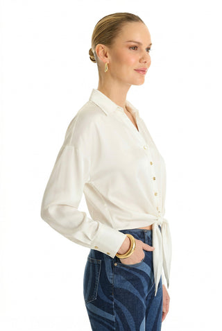 A woman stands in profile against a plain white background, wearing the Cecilia Silk Tie-Front Blouse in white with a collar, styled with blue patterned jeans, gold hoop earrings, gold bracelets, and her hair neatly pulled back.