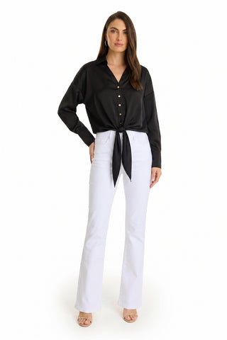 A woman faces forward wearing the Cecilia Silk Tie-Front Blouse in black, paired with white high-waisted flare jeans, nude heels, and hoop earrings against a plain white background.