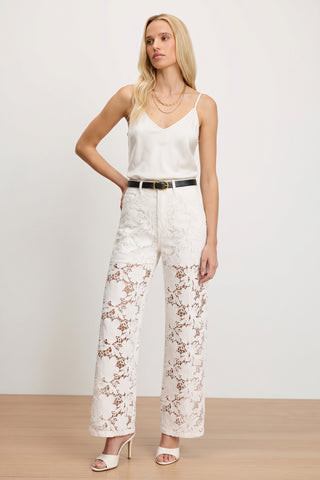 A woman stands against a plain background wearing a white satin camisole, the Celeste High Rise Jean – White styled as high-waisted lace pants with a black belt, and white heeled sandals. She has long blonde hair and is accessorized with layered gold necklaces.
