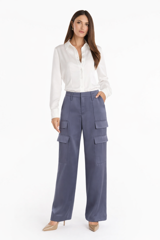 A woman with straight brown hair and a neutral expression wears a white blouse, beige pointed-toe heels, and the Milan Satin Cargo Pant in navy—a high-waisted satin-style trouser with multiple pockets—against a white background.