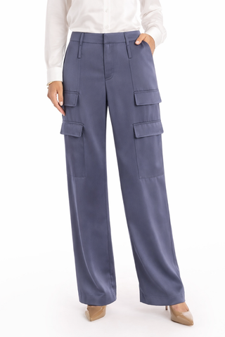 A person stands on a plain white background wearing the Milan Satin Cargo Pant in navy with multiple pockets, paired with a white button-down shirt and beige pointed-toe shoes, one hand casually in a pocket.