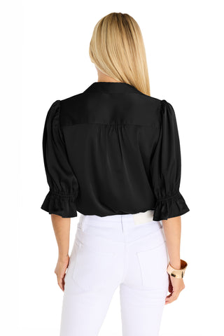 A woman with long blonde hair is seen from the back wearing the Cindy Ruffle Sleeve Shirt in black and white pants, accessorized with a tan bracelet, standing against a plain white background.