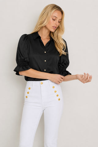 A woman with long blonde hair wears the Cindy Ruffle Sleeve Shirt in black, featuring gold buttons, paired with high-waisted white pants with gold buttons, standing against a neutral background.