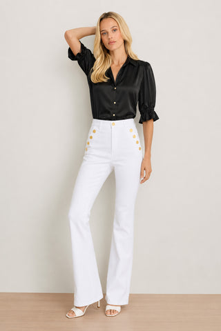 A woman stands against a light background, wearing the Cindy Ruffle Sleeve Shirt in black with puff sleeves, paired with high-waisted white flared pants featuring gold buttons and white heeled sandals. She has blonde hair and poses with one hand touching her head.