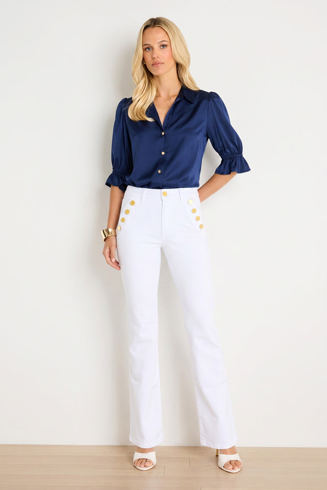 A woman stands against a plain light wall, wearing the Cindy Ruffle Sleeve Blouse in navy with white high-waisted pants featuring gold buttons. She completes the look with white heels, a gold bracelet, and her long blonde hair worn down.