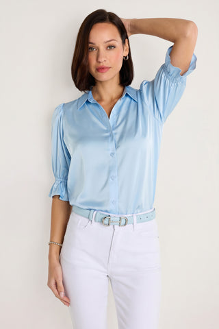 A woman with short brown hair and a neutral expression wears the Cindy Ruffle Sleeve Blouse in sky blue with elbow-length sleeves and white pants, standing against a plain background with one arm raised to her head.