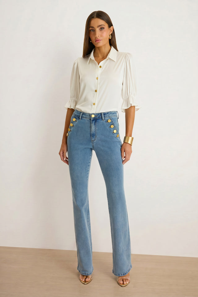 A woman stands against a plain background wearing the Cindy Ruffle Sleeve Blouse in white, styled with high-waisted blue jeans featuring gold button details, a gold bangle, and open-toe heels.