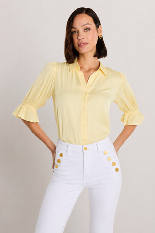 A woman stands with hands on hips against a plain background, wearing the Cindy Ruffle Sleeve Shirt in sunshine yellow and white high-waisted pants with gold buttons.