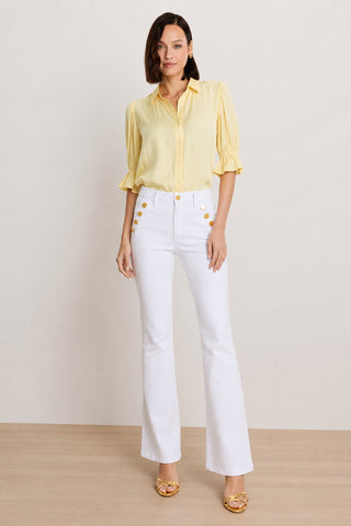 A woman with shoulder-length brown hair and gold hoop earrings poses against a plain background in the Cindy Ruffle Sleeve Shirt in sunshine yellow, paired with white high-waisted pants with gold buttons and yellow heeled sandals.