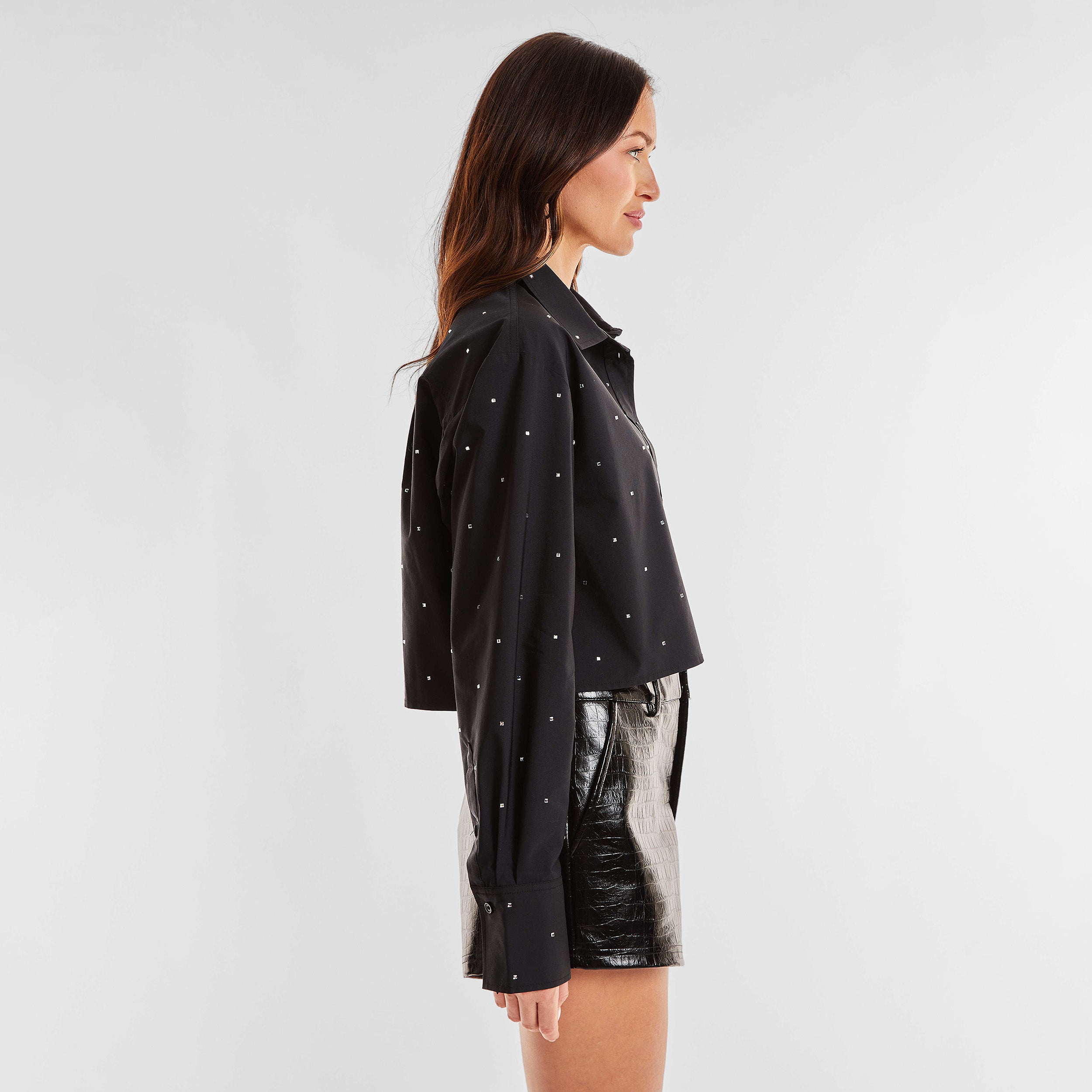 Side view of woman wearing a black cropped button down shirt with crystal embellishments and croco pattern embossed faux leather shorts.