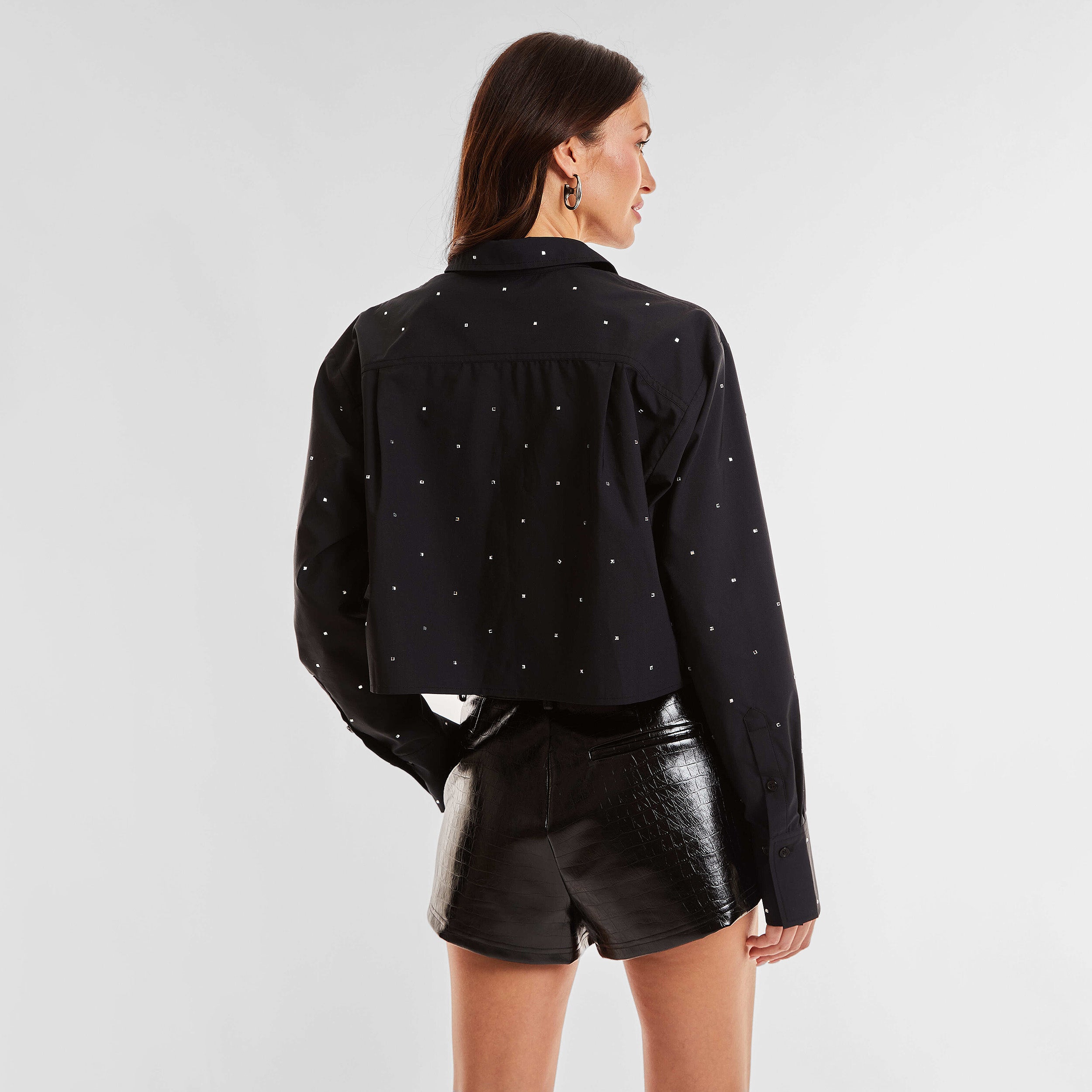Rear view of woman wearing a black cropped button down shirt with crystal embellishments and croco pattern embossed faux leather shorts.