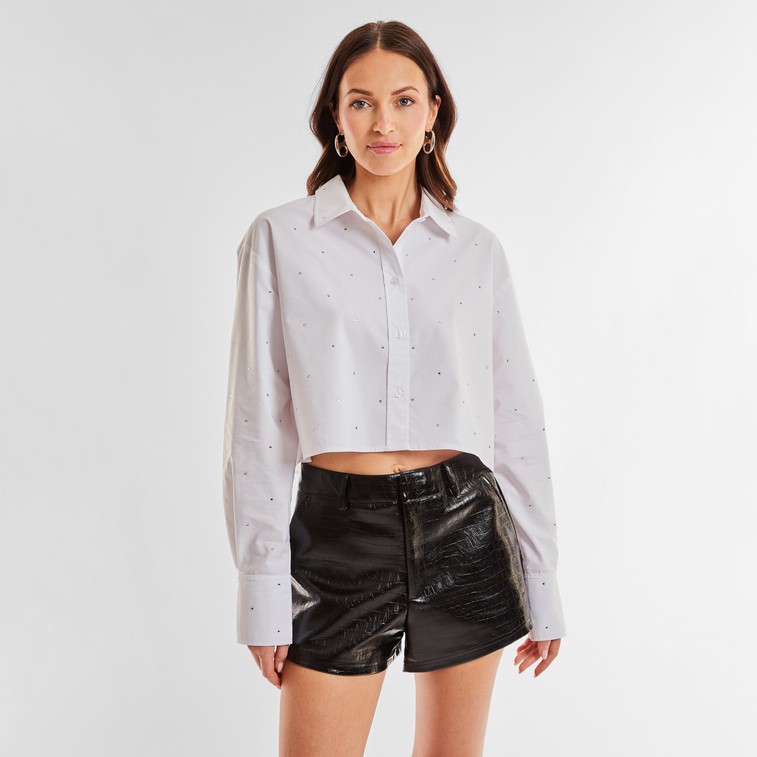 Front view of woman wearing a white cropped button down shirt with crystal embellishments and black croco pattern embossed faux leather short.