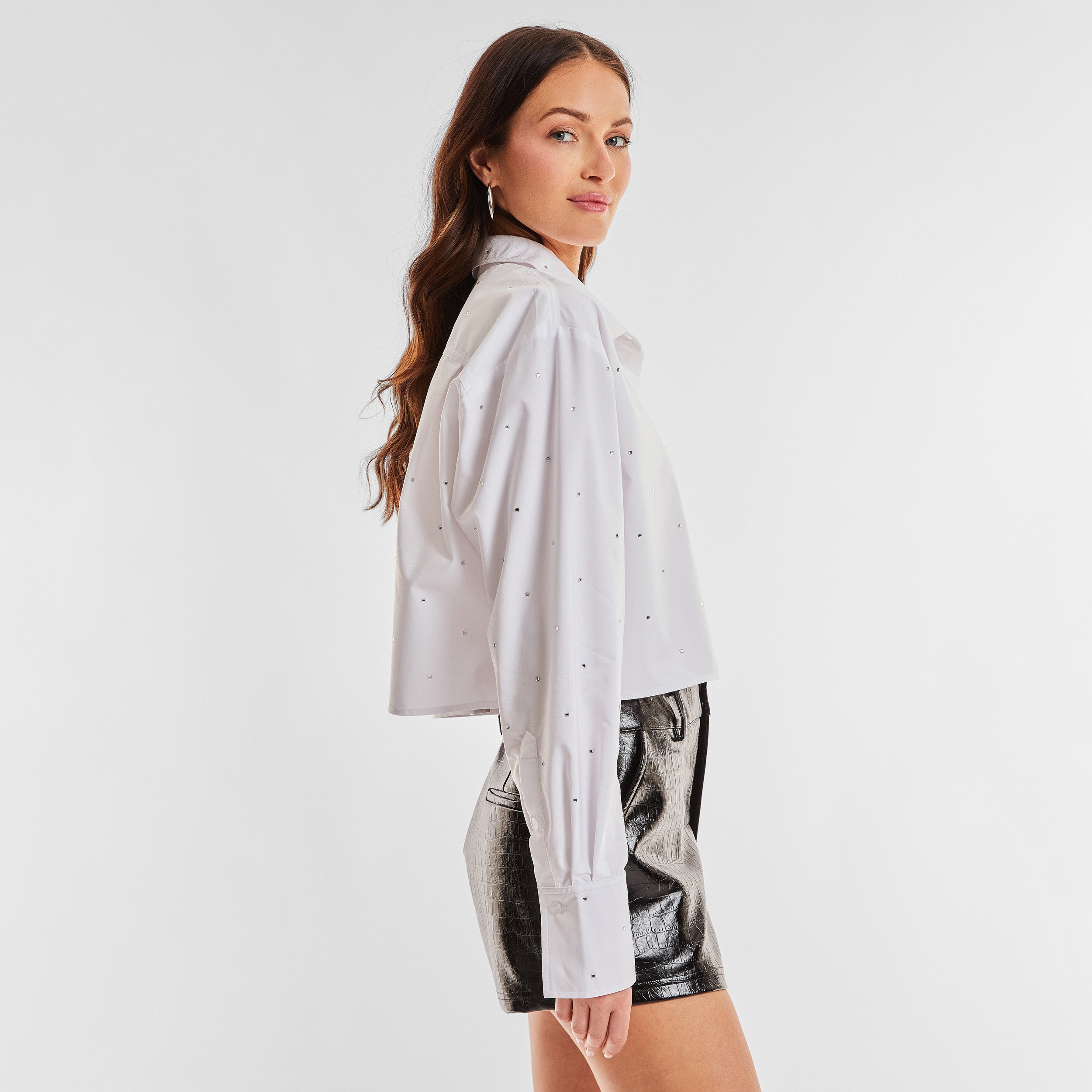 Side view of woman wearing a white cropped button down shirt with crystal embellishments and black croco pattern embossed faux leather short.