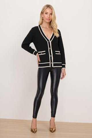 A woman stands against a plain background wearing the Devin Rose Contrast Cardigan in black, paired with shiny black leggings and gold high heels.