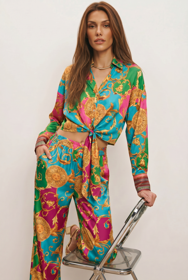 A woman poses in the Cecilia Silk Tie-Front Blouse - Dolce Sun, a vibrant vacation top with colorful patterns and a knotted front, paired with high-waisted pants. She leans on a clear chair, her long hair flowing gracefully.