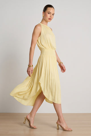 A woman wears the Emelia Halter Dress in Sunshine Yellow with clear heeled sandals, gold hoop earrings, and gold bracelets as she walks across a light wooden floor against a plain, light background.
