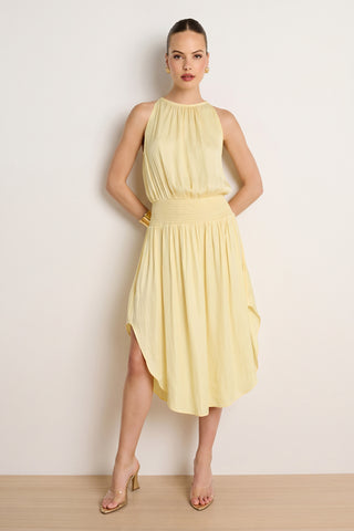 A woman stands against a plain wall wearing the Emelia Halter Dress in sunshine yellow, featuring a sleeveless design, gathered waist, and asymmetrical hem, styled with clear-heeled sandals and gold accessories.